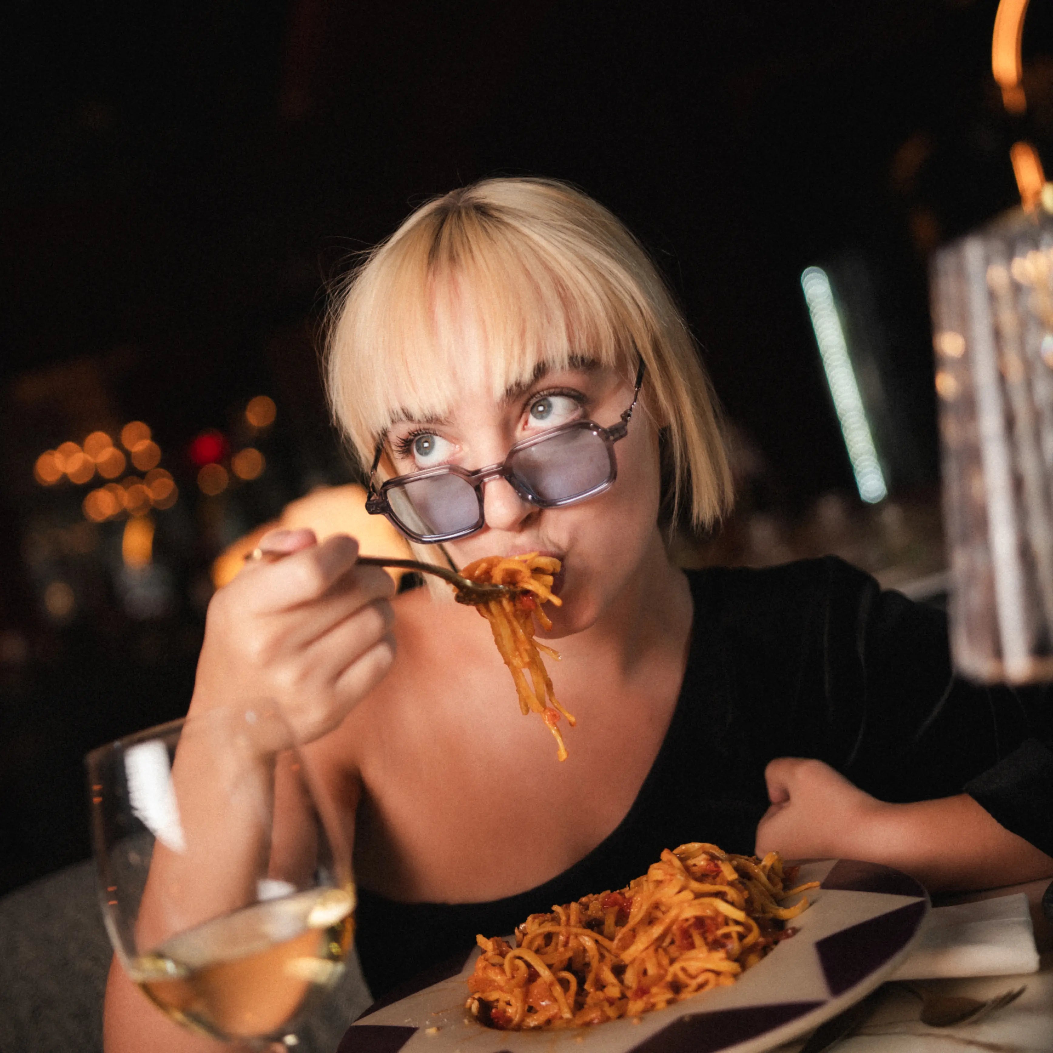 A woman wearing Bold Silver Sunglasses enjoying pasta in a restaurant setting, with elegant decor and a glass of wine.